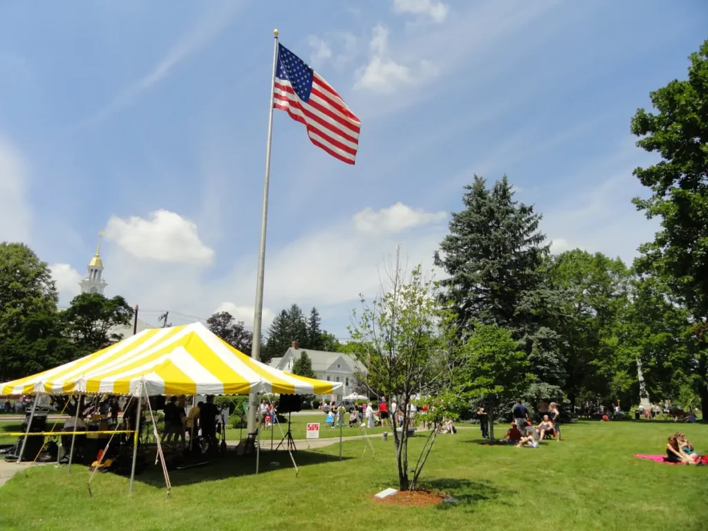 Billerica Town Common on a summer day, with a flag, a striped tent and neighbors gathered on the lawn